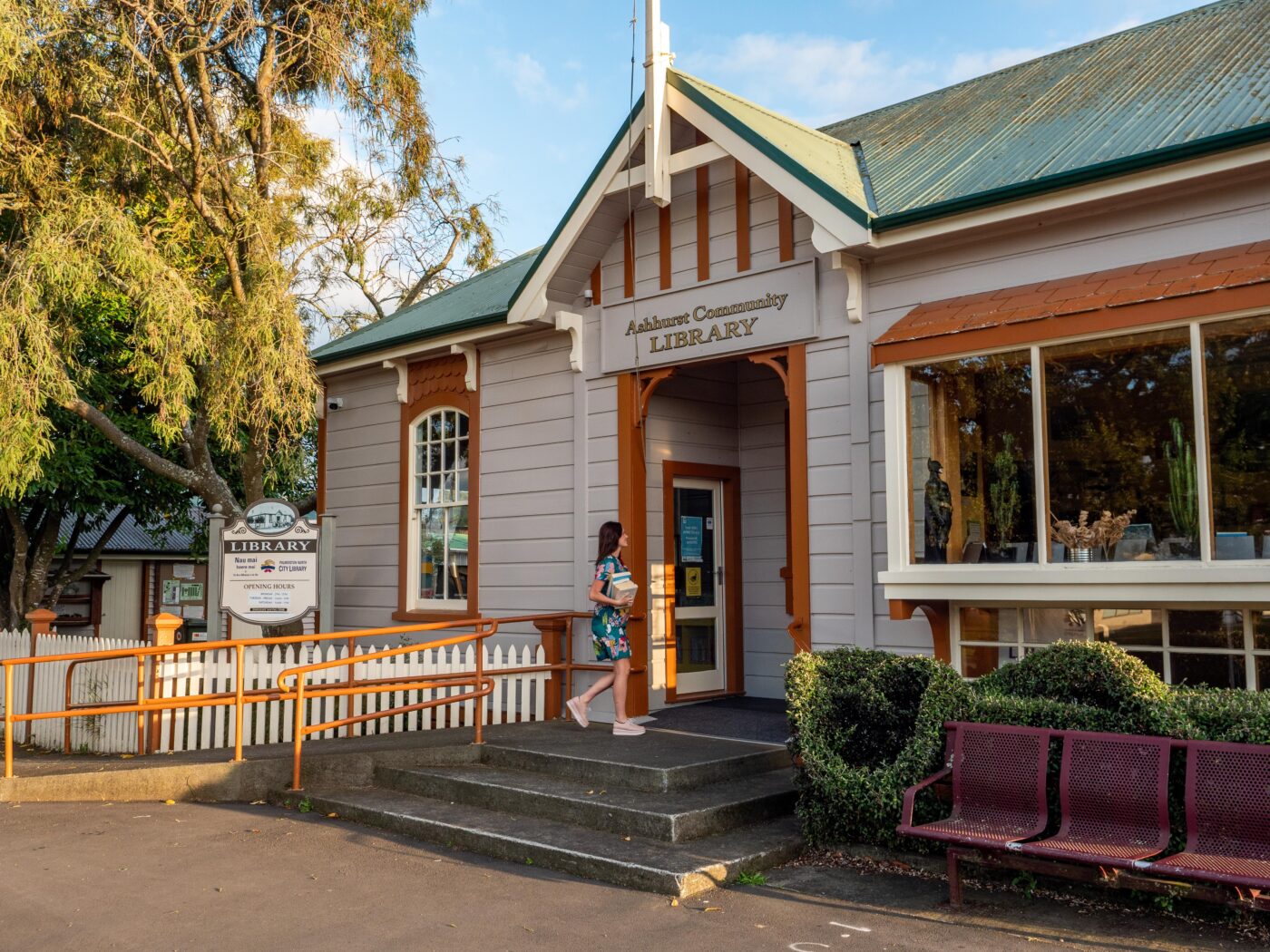 A woman walking up to a grey library building with orange trim and green roof and a white picket fence.