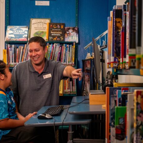 Librarian sitting with a child and pointing at a computer screen in a corner surrounded by books.