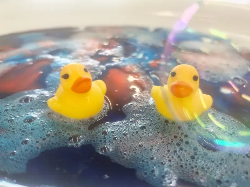 Two yellow rubber ducks are sitting in colourful slime. They're surround by bubbles.
