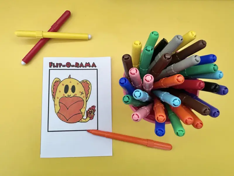 Felt pens are scattered across a yellow table beside a child's animal drawing