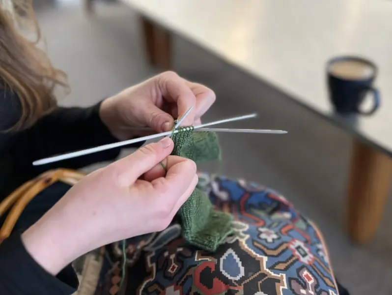 Close-up of a person's hands knitting a pair of green gloves, with a cup of coffee on a table in the background