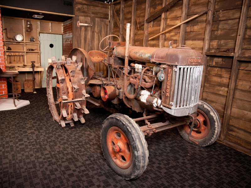 Photograph of a rusted old tractor in a carpeted room, next to a wall of unpainted wooden boards. The tractor appears dusted with old mud and has large, spikily ridged back wheels. Behind the tractor is an alcove with shelves holding antique objects.
