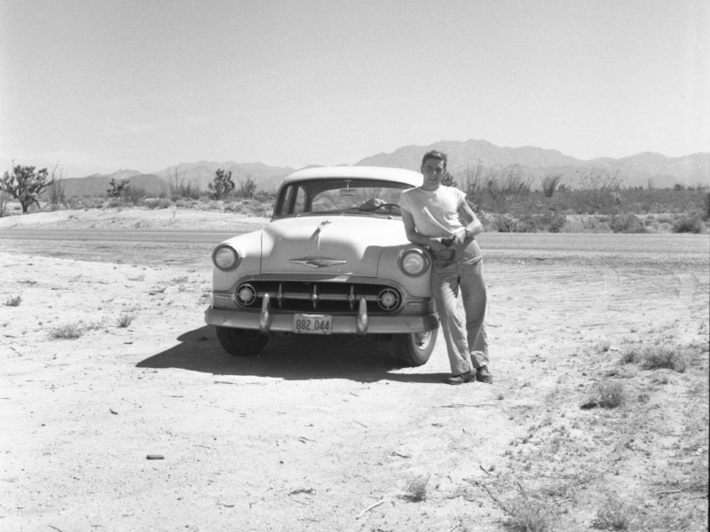 Black-and-white photograph of a man leaning against the hood of a car in a desert landscape, smiling into camera. Behind the car, a road bisects the frame, and behind that is a bank of scrubby plants and a range of mountains.