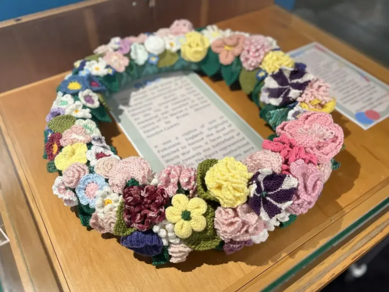 A colourful wreath of knitted flowers with text attributing its construction to the library's Heritage Craft Group.