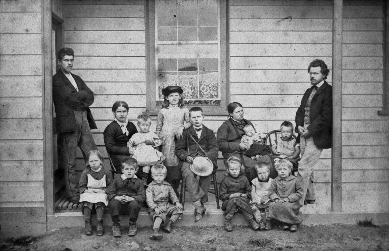 Black and white photo of a family on a porch. Two men in black jackets flank the women and children seated with one man in the centre holding a hat in his hand.