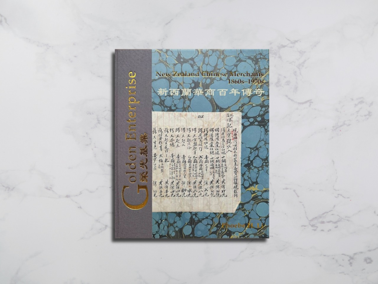 Book with grey spine and gold lettering. The cover has an image of a piece of paper with Chinese with a blue and gold marbleized background.