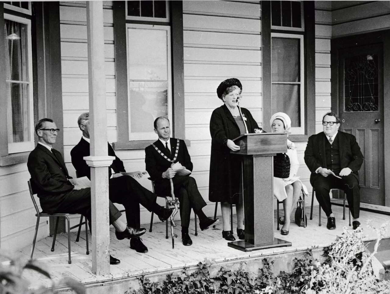 Julia Wallace, a short woman with black coat and black round hat speaking at a lecturn on a front porch. Three men, one with mayoral chains are seated to her left and a woman in white and a man are seated to her left.