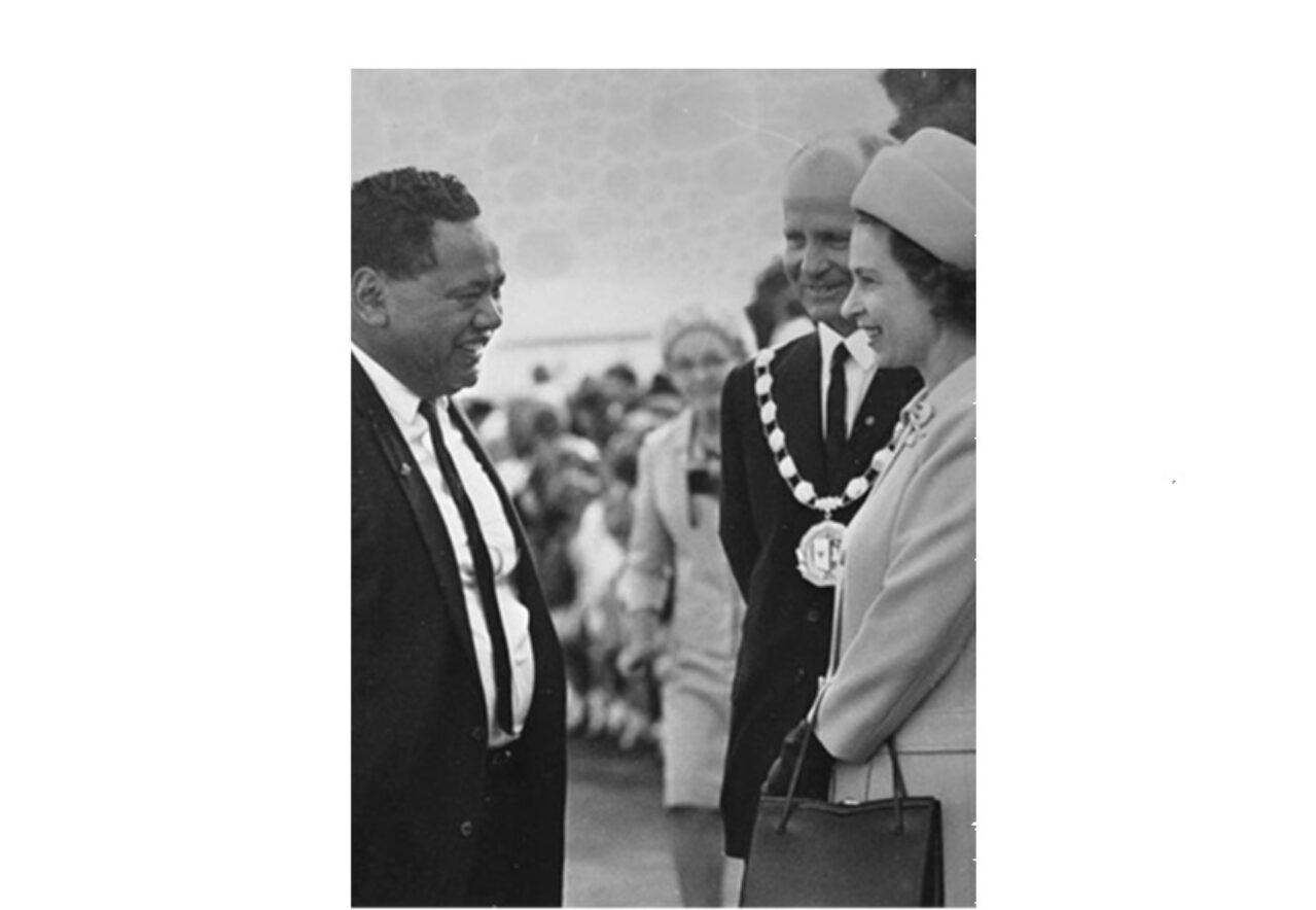 Black and white photo of a man in a suit (Teira Samuel Mihaere) smiling and meeting with Queen Elizabeth II and Prince Phillip. Queen Elizabeth is wearing a hat that matches her coat and holding a handbag. Prince Philipp has gold chain around his neck.