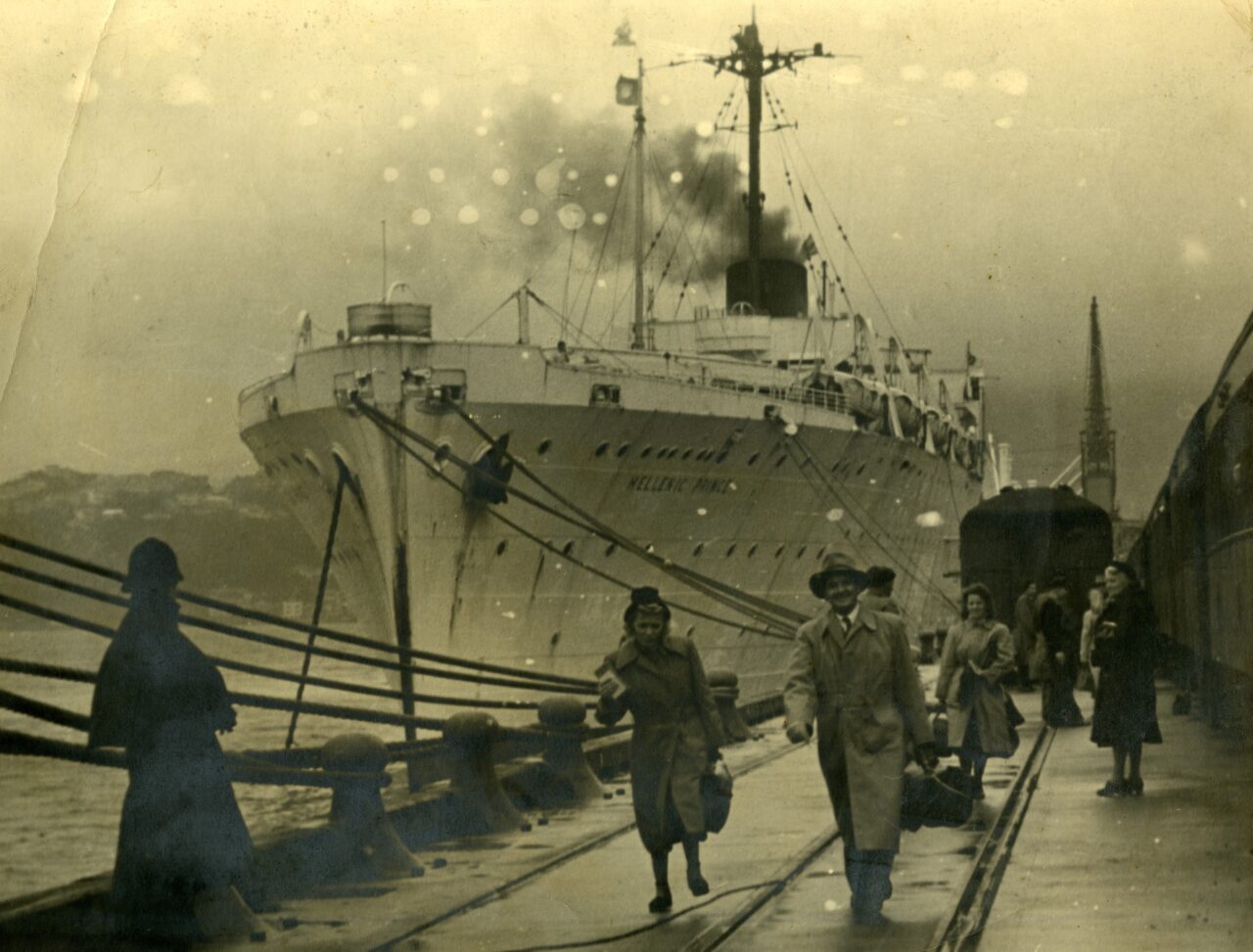 Sepia image of a steamer ship docked. People with luggage are walking up the dock from the ship.