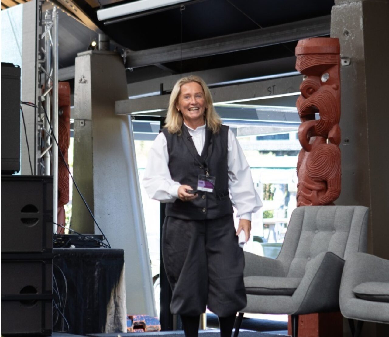 Woman wearing a white blouse and black v-neck dress and black boots smiling walking towards the camera. There is a Māori Pou to the right of the woman.