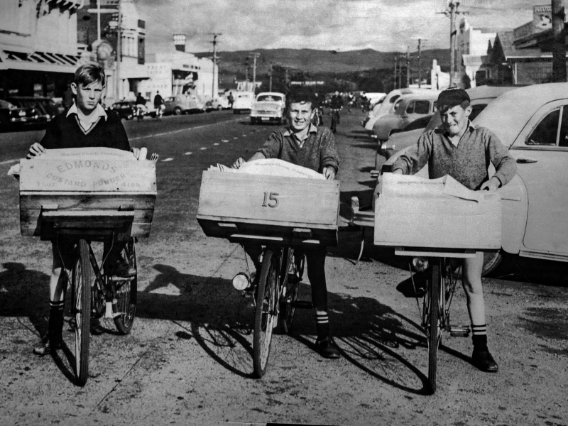 A black-and-white photograph of 3 teenaged boys standing with bicycles on a street. The bicycles are mounted with wooden crates filled with newspapers. There are buildings, parked cars and more cyclists in the background, and hills on the horizon.
