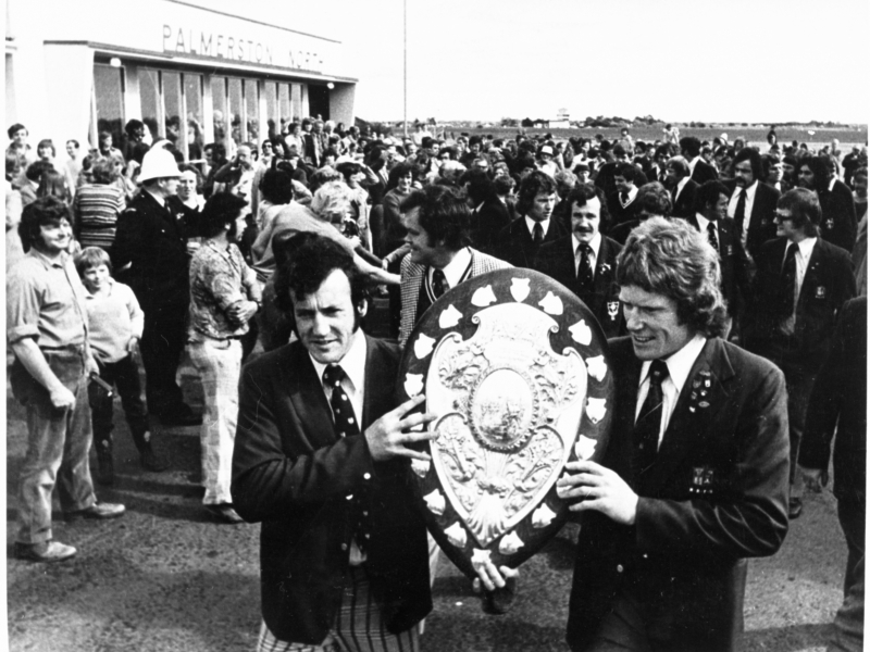 Black-and-white photograph of 2 men holding up a decorated shield, with a large crowd behind them. The crowd are gathered outside a single-storey building with the words "Palmerston North" on its façade.