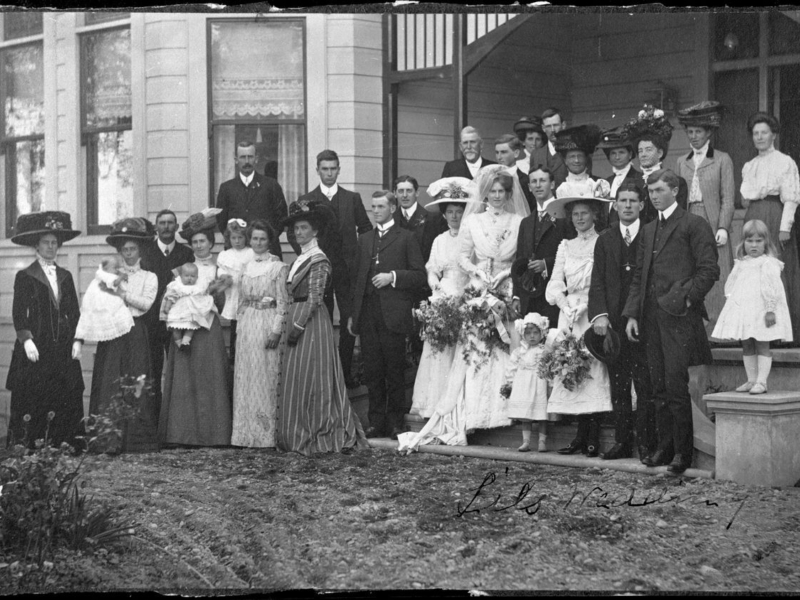 Black-and-white photograph of a wedding party posed outside a house, dressed in Edwardian formal attire.