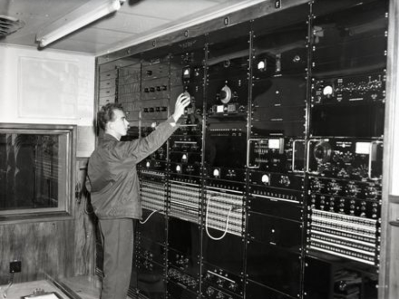 Black and white photo of a man working along a wall array of various radio equipment, dials, switches and connections.