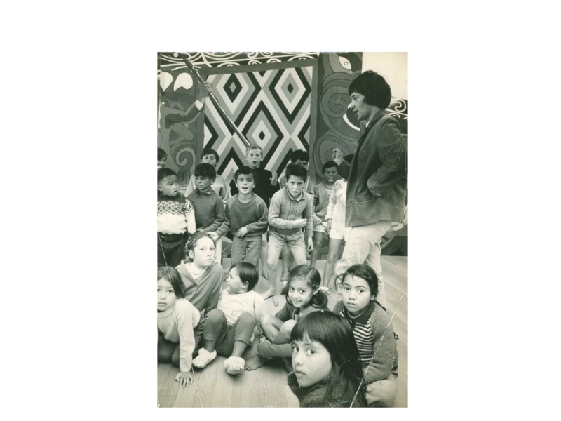 A woman standing speaks to a group of children who are standing and sitting. There is a mural in the background.