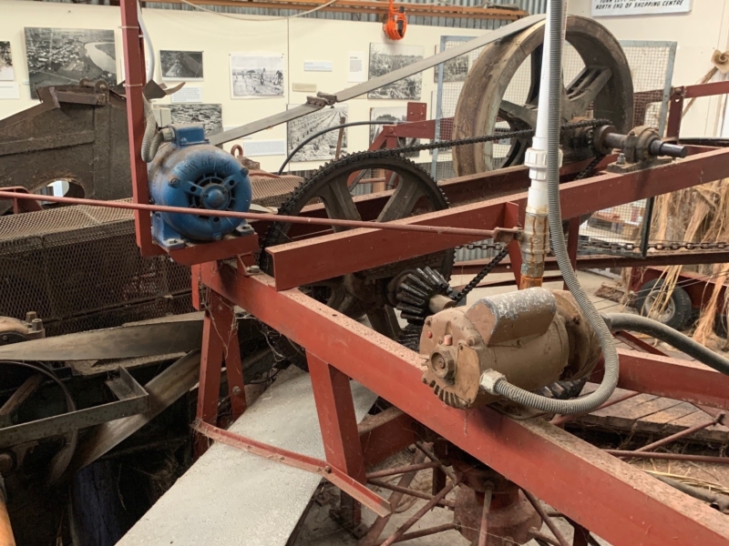 Photo of a room with various industrial machines wheels and belts. The machine in the foreground has red bars a blue engine and rotating wheels.