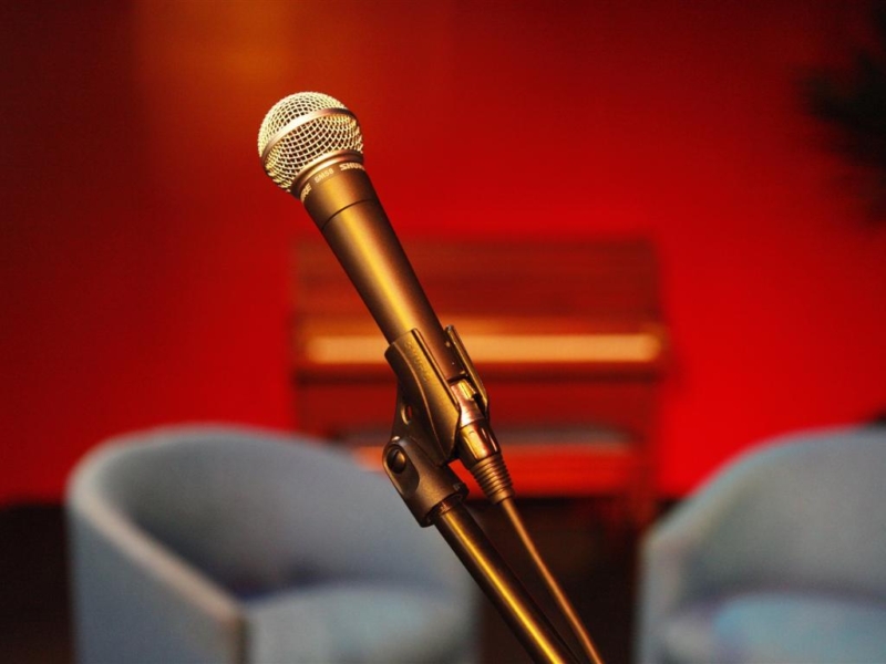 Black microphone in front of two blue chairs, a piano and a red wall.