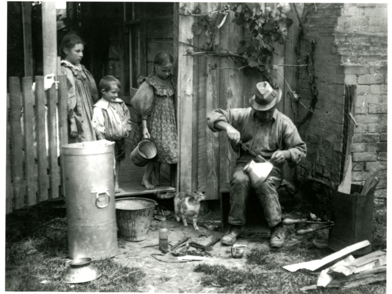 Black and white photo of man sitting with tools around him, working on a metal cylinder with three children looking on from a wooden porch.