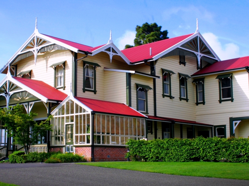 Photo of a stately yellow house with green and white trim, red roof and glass conservatory surrounded by lush green hedge trees and plantings.