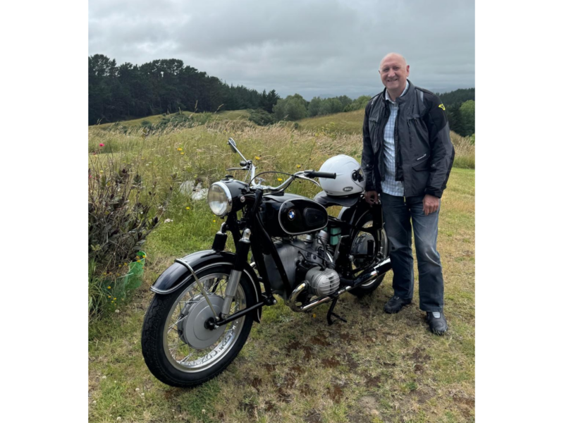 Photograph of a man standing beside a motorcycle and smiling into camera, against a background of rolling hills.