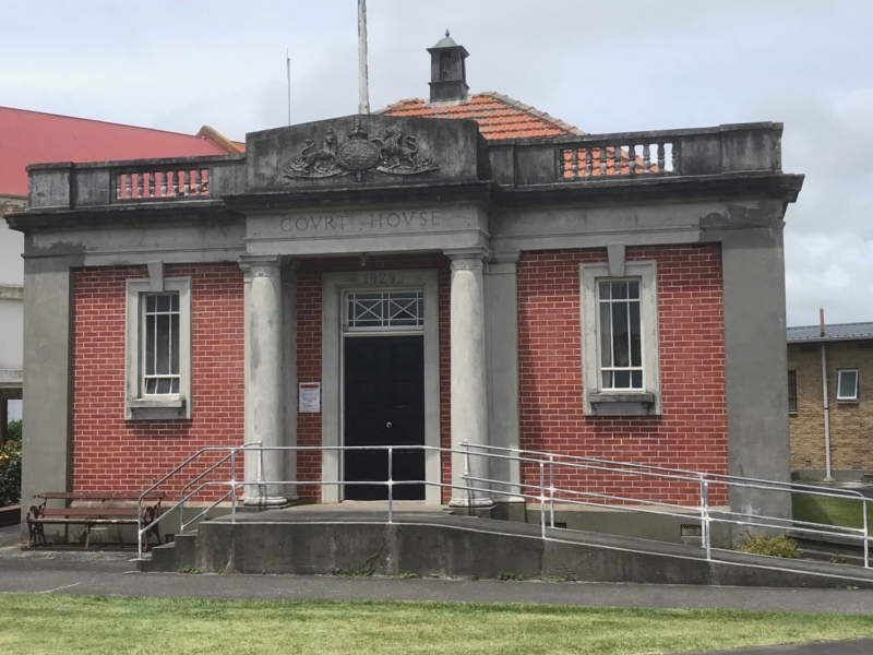 Photo of red brick and concrete building with two ionic columns saddling the black door. The windows have keystones and have vertical bars over the windows. The pediment is carved and has the word courthouse.