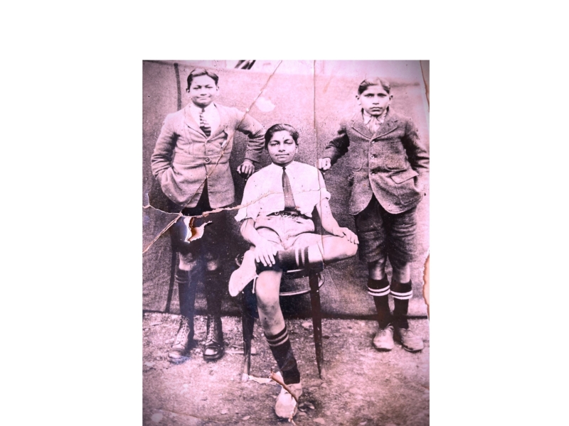 Pink tinted image with three boys, one sitting in a chair with a white shirt, black tie shorts and long socks. The two other boys are in suit jackets shorts and long socks behind.