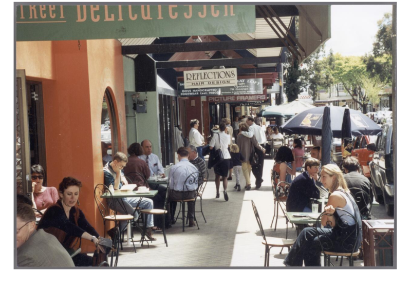 Colour photo of people sitting at cafe tables along a sidewalk. There is an orange wall to the left, underside of verandas and shop signs hanging from the verandas.