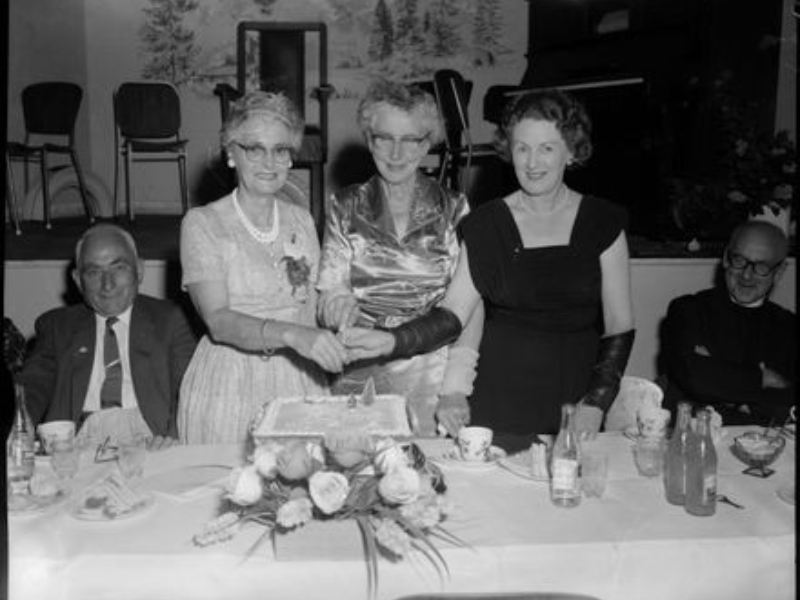 A black and white photo of three women in party dresses holding a knife over a fancy cake. There are two men sitting one to the left and one to the right, looking on.