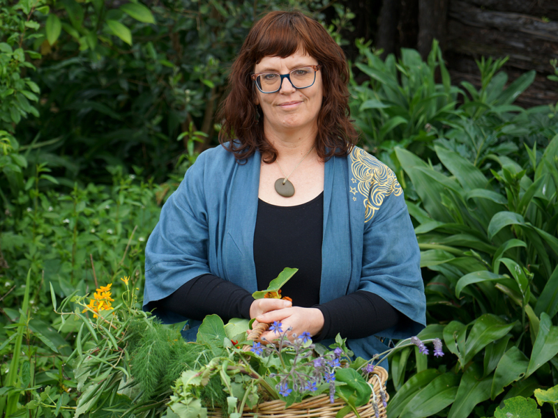Photograph of a smiling woman wearing a blue shawl and holding a wicker basket full of herbs and flowers. She is surrounded by tall, green undergrowth.