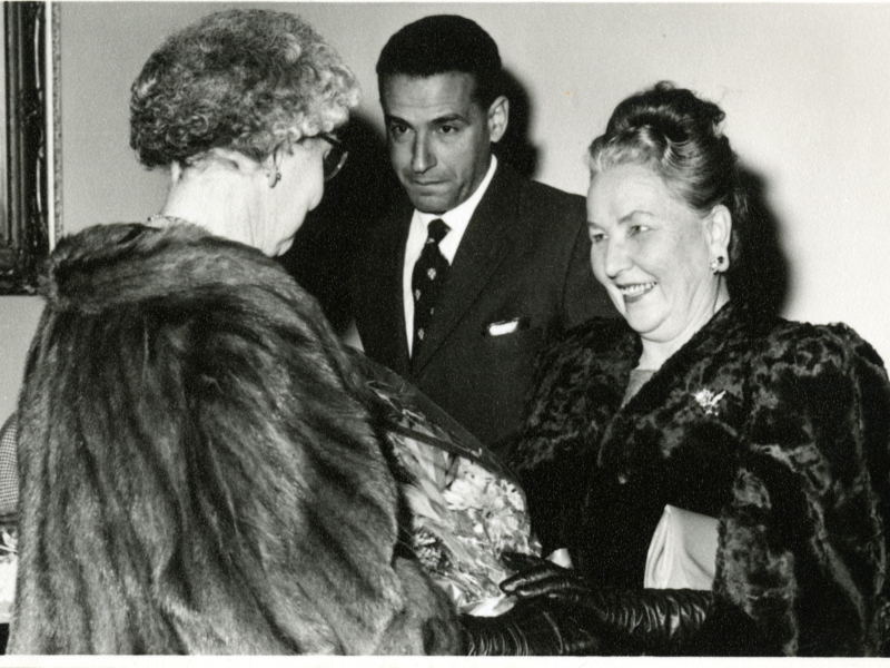 Black and white image of two women in fur coats greeting each other. There is a man with a suit and tie in the background.