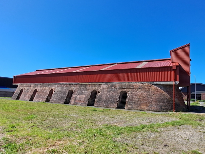 Coloured photo of long building in a green grass field. Brick base with arched entrances and red tin roof with tower.