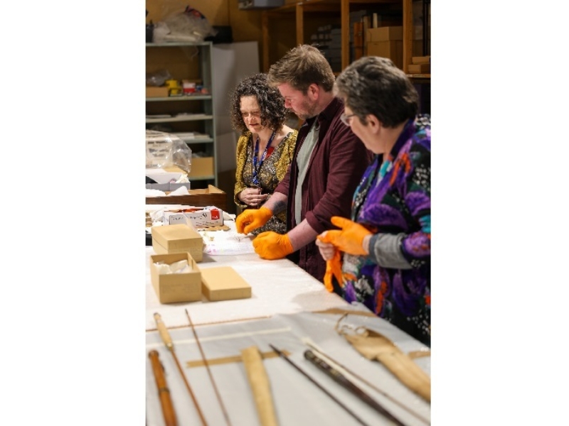 Photo of three people with orange gloves inspecting a table of boxes and museum artefacts.