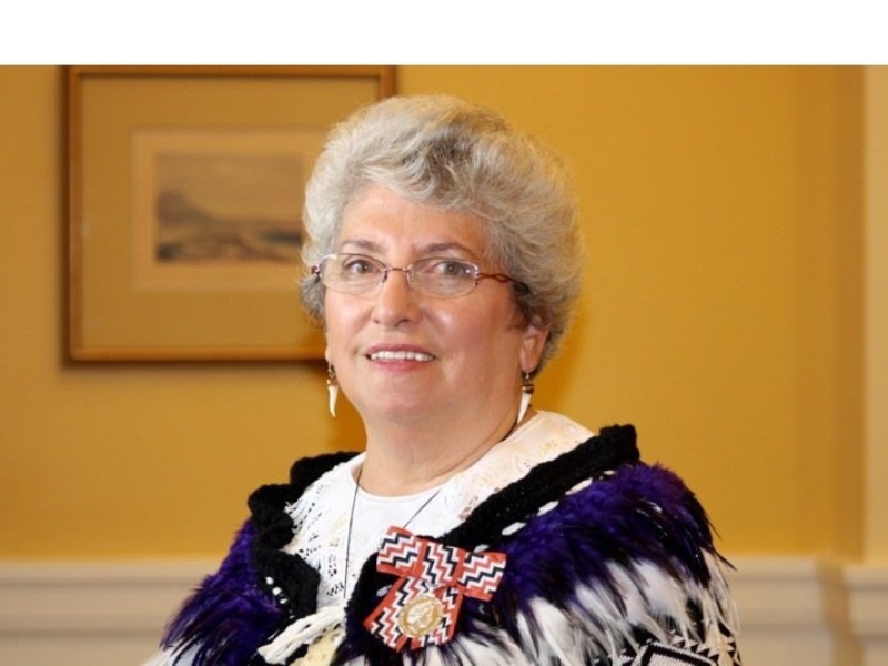 Woman with glasses, short grey hair, black, purple and white kākahu, and a badge with a red, white and black poutama design. She is standing in front of a yellow wall with a framed picture.