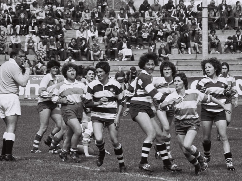 Black and white photo of women in striped shirts playing rugby at a stadium with a large crowd in the background.