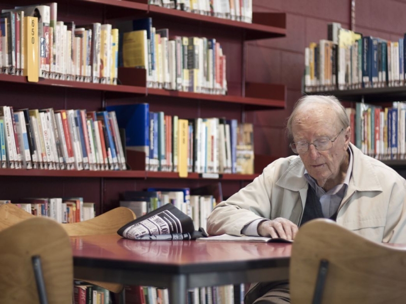 Merv Hancock in a khaki jacket sitting at a table reading surrounded by red library bookshelves.