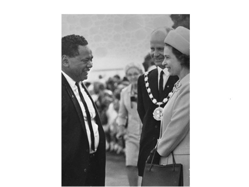 Black and white photo of a man in a suit (Teira Samuel Mihaere) smiling and meeting with Queen Elizabeth II and Prince Phillip. Queen Elizabeth is wearing a hat that matches her coat and holding a handbag. Prince Philipp has gold chain around his neck.