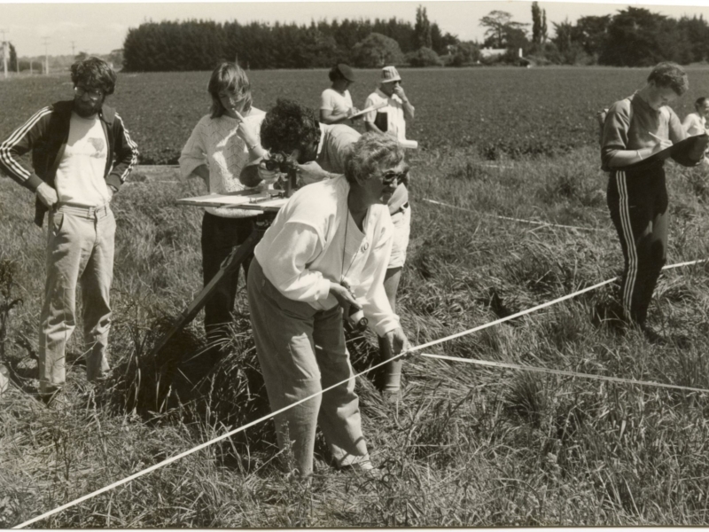 Black and white photo of a woman (Mina McKenzie) in white blouse and grey pants holding ropes strung across a field. There is a man with a surveying tool behind her and five other people standing overlooking.