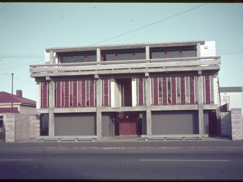 Streetscape photo of a concrete architecturally designed hall with whakairo (Māori carvings) in wood vertically along the first floor.