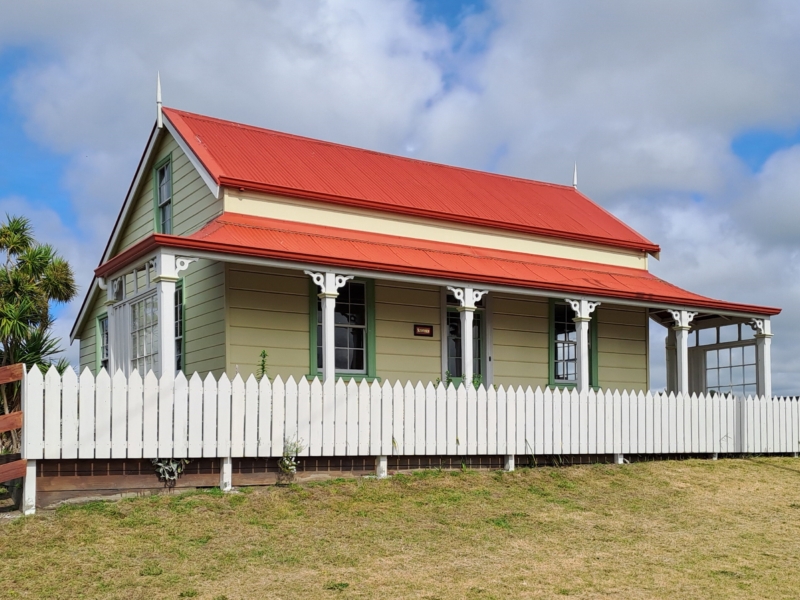 Yellow wooden cottage with red roof, green window frames and green picket fence. Cabbage tree to the left and blue skies with fluffy clouds above.