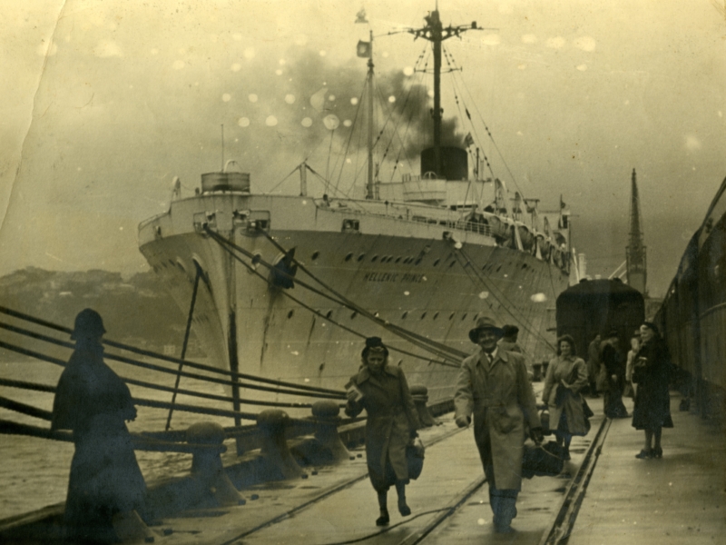 Sepia image of a steamer ship docked. People with luggage are walking up the dock from the ship.