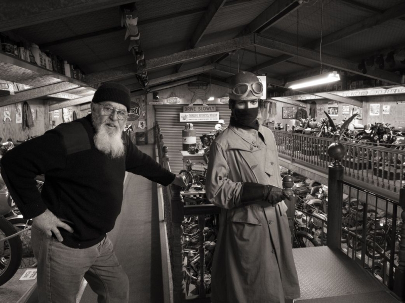 Man in black jumper and black beanie with white beard standing by a mannequin dressed in an army jacket. In the background are hundreds of motorcycles.