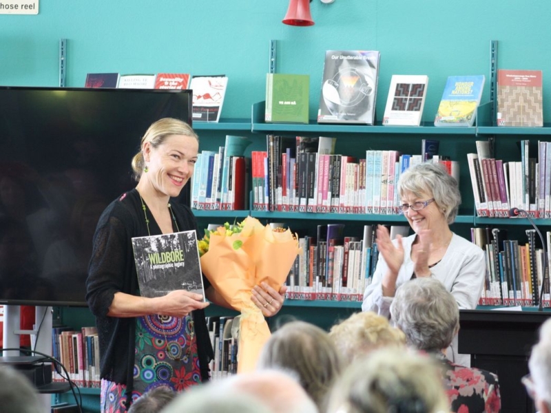 Catherine Knight, blond hair back in a pony tail, standing holding her book and a bunch of flowers in an orange wrap. Margaret Tennant in a white suit jacket looks on about to clap.