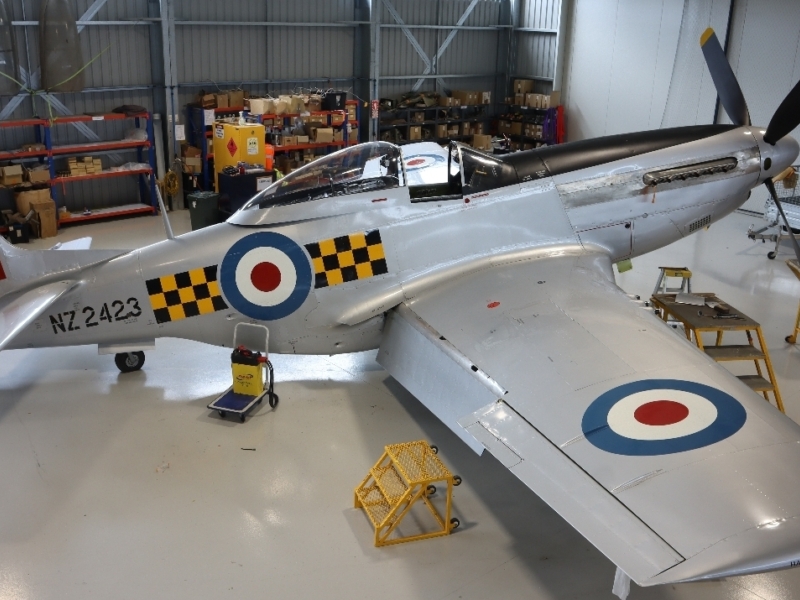 Silver airplane in a hanger being worked on. The airplane has a black propellor with yellow tips, and blue, white and red bullseyes on the wings and side. There is also black and yellow checks.