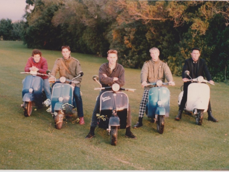 Six young men on vespa style scooters in an arrow formation on the lawn with trees in the background.