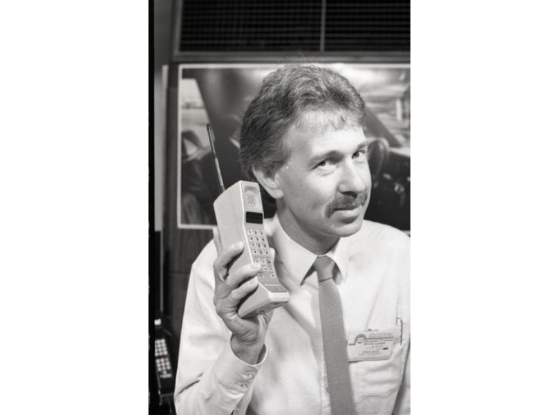 Man in a white shirt and tie, holding an early cellular phone.