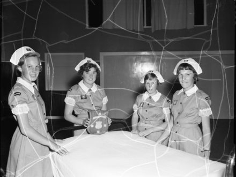 Four young women in St. John nursing uniforms hold a plaque award over a hospital bed.