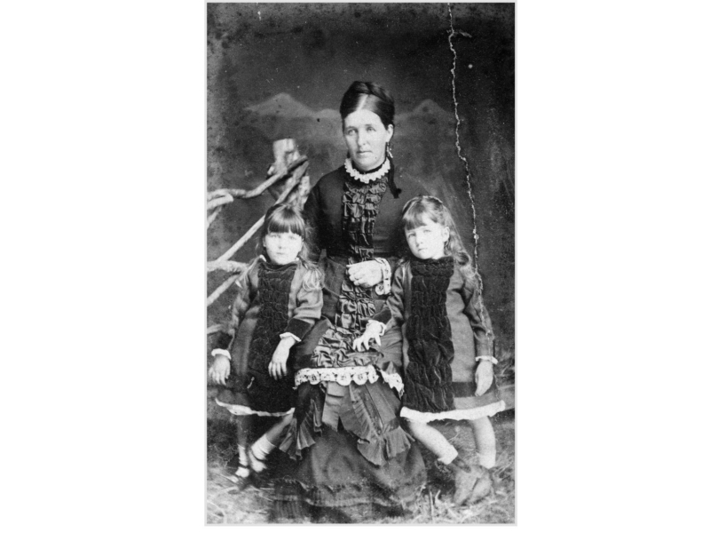 Black and white portrait of a woman with two daughters. All are wearing dresses with black velvet strip down the front.