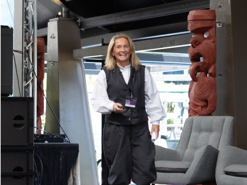 Woman wearing a white blouse and black v-neck dress and black boots smiling walking towards the camera. There is a Māori Pou to the right of the woman.