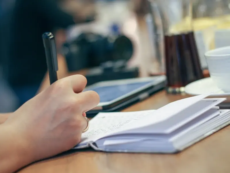 Close-up photograph of a hand holding a pen, resting on a table that holds a notebook, a tablet, a camera and cups and saucers.