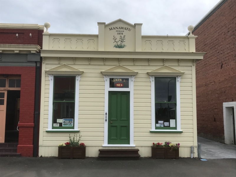 Photo of a yellow building flanked by two brick buildings. The building has white trim around two windows either side of a green door. Prominent pediments over the door and windows. Manawatu Herald 1878 with flax painted on the cornice. Main St. No. 6 above the door.